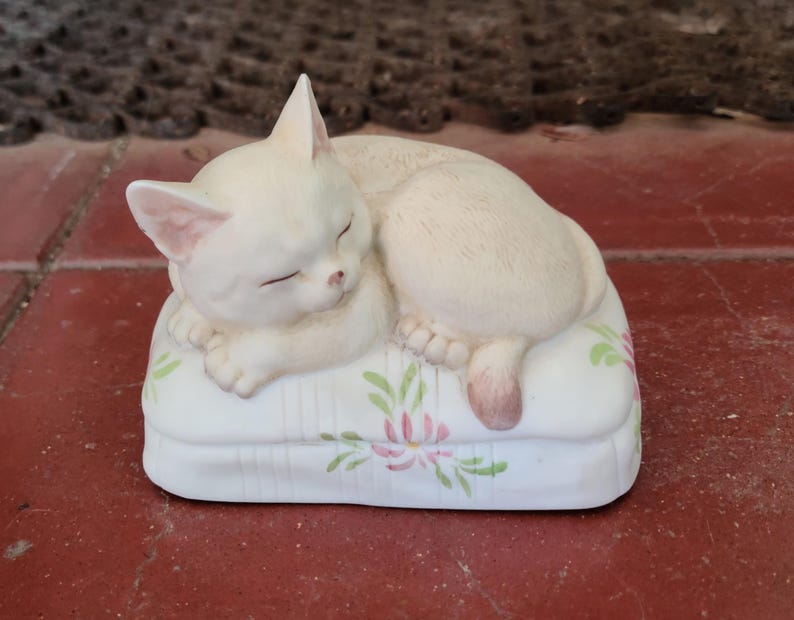 May include: A cream-colored ceramic cat figurine sleeping on a white pillow. The pillow is decorated with pink and green floral designs. The cat is curled up, with its eyes closed and its tail resting on the pillow.