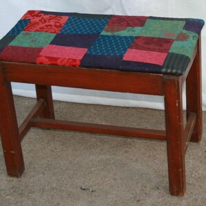 May include: A wooden stool with a patchwork fabric seat in shades of red, green, blue, and black. The stool has a simple design with four legs and a single crossbar.