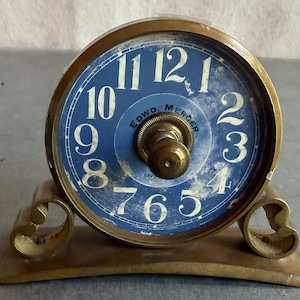 May include: A round brass desk clock with a blue face featuring white numerals from 1 to 12. The center of the clock displays the text EDWD. MERCER in black lettering. The clock is mounted on a curved brass base with decorative scroll feet.