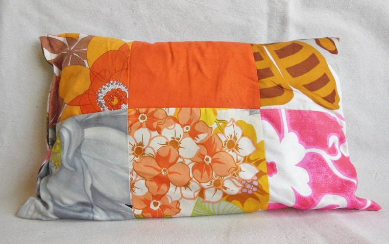 May include: Rectangular patchwork pillow featuring a vibrant mix of floral patterns in orange, yellow, pink, and gray. The pillow's design includes large flower motifs and solid orange squares, creating a retro, bohemian aesthetic. A decorative accent for a bed or sofa.