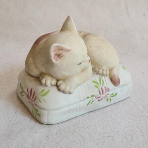 May include: A cream-colored ceramic cat figurine sleeping on a white pillow. The pillow has a floral design in pink and green. The cat is curled up with its eyes closed, and its tail is visible.
