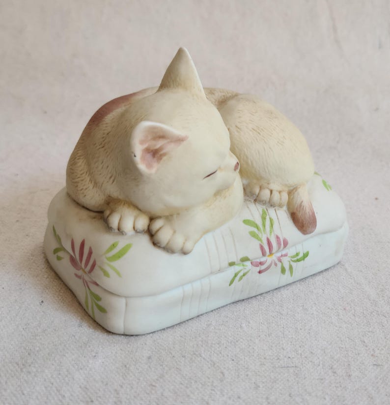 May include: A cream-colored ceramic cat figurine sleeping on a white pillow. The pillow has a floral design in pink and green. The cat is curled up with its eyes closed, and its tail is visible.