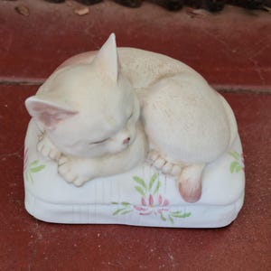 May include: A cream-colored ceramic cat figurine curled up asleep on a white pillow. The pillow is decorated with pink and green floral designs. The cat's eyes are closed, and its paws are tucked in.