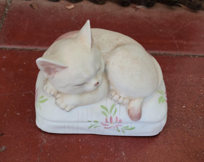 May include: A cream-colored ceramic cat figurine curled up asleep on a white pillow. The pillow is decorated with pink and green floral designs. The cat's eyes are closed, and its paws are tucked in.
