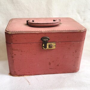 May include: A vintage, rectangular, pink makeup case with a textured exterior. The case has a brown handle and a metal clasp with a keyhole. The case appears to be made of a durable material, and shows signs of wear.