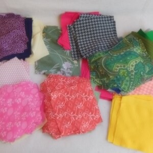 May include: A collection of fabric scraps in various colors and patterns, including pink, green, yellow, orange, and purple. The fabric scraps are arranged in a pile on a white surface.