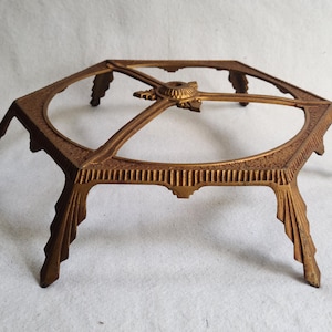 May include: A vintage, ornate, bronze-colored metal stand with an octagonal top and six angled legs. The top features a circular opening and decorative detailing. The legs have a stylized, fan-like design. The stand is likely for decorative purposes.