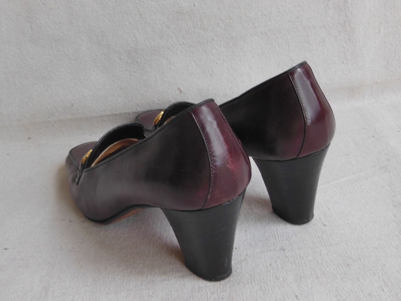 May include: A pair of burgundy leather heeled shoes with a gold buckle detail. The shoes have a black heel and a pointed toe.