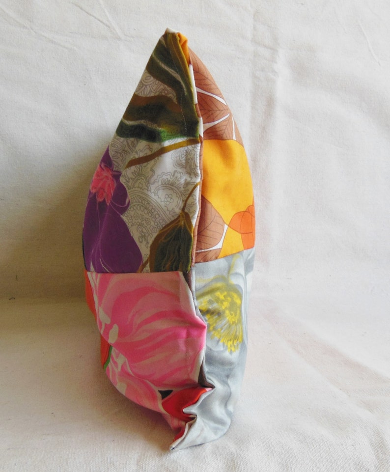 May include: A patchwork pillow with a variety of floral patterns in shades of pink, purple, orange, and gray. The pillow is rectangular and appears to be made of fabric scraps sewn together. The pillow is on a white surface.