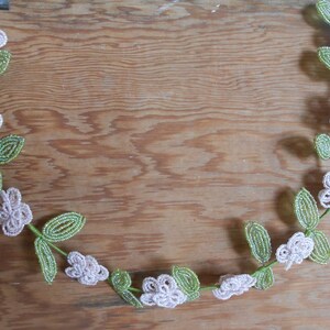 May include: A beaded garland with white flowers and green leaves. The garland is made of small beads and is strung together to form a continuous loop.
