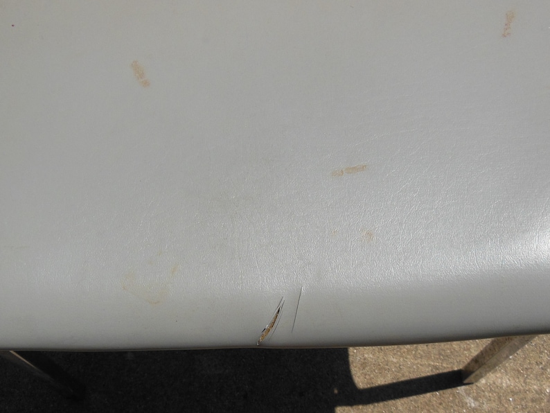 May include: A close-up of a gray chair seat with a small tear and some light brown stains.