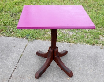 May include: A square table with a bright magenta painted top and a dark brown wooden pedestal base. The table has four legs that extend from the base.