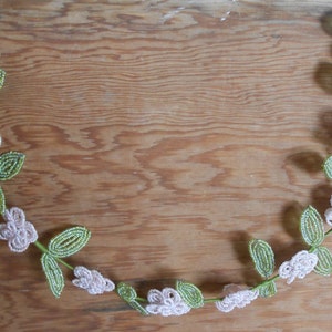 May include: A beaded garland with white flowers and green leaves. The garland is in the shape of a crescent moon and is hanging on a wooden surface.