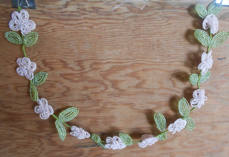 May include: A beaded garland with white flowers and green leaves. The garland is in the shape of a crescent moon and is hanging on a wooden surface.