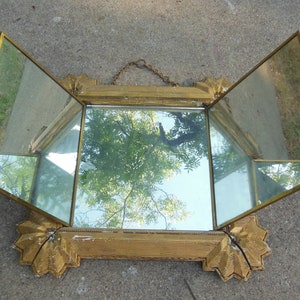 May include: A gold-toned, ornate, three-sided, hinged mirror with a chain for hanging. The mirror is reflecting a tree and sky.