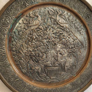 May include: An antique metal plate with an intricate embossed design. The plate features birds, flowers, and foliage, with a detailed border. The color is a mix of silver and bronze, with some areas showing a green patina.