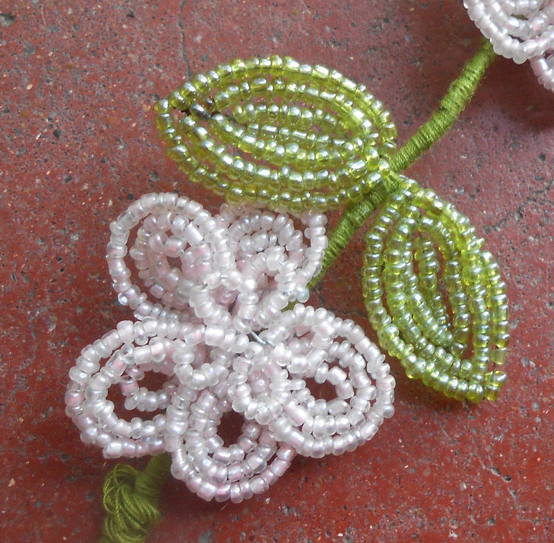 May include: A beaded flower with five petals in shades of white and pink, with two green leaves. The flower and leaves are made of small, round beads.