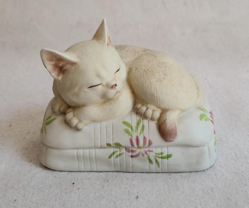 May include: A cream-colored ceramic cat figurine sleeping on a white pillow. The pillow has a floral design with pink and green accents. The cat's eyes are closed, and its paws are tucked in. The figurine is a decorative item.