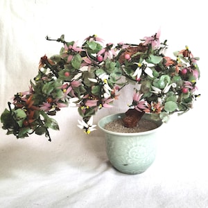 May include: A decorative bonsai tree with pink, white, and green leaves and flowers. The tree is in a light green ceramic pot with a detailed floral design. The bonsai has a brown trunk and is set against a white background.