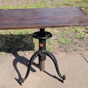 May include: A vintage wooden table with a black metal base. The table has a crank to adjust the height. The table is on a concrete surface.