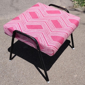 May include: A pink and white patterned footstool with a black metal frame. The footstool has a geometric design and is perfect for adding a touch of color to any room.