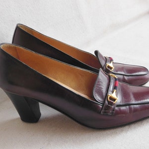 May include: A pair of burgundy leather heeled shoes with a gold buckle and a red and blue stripe detail.