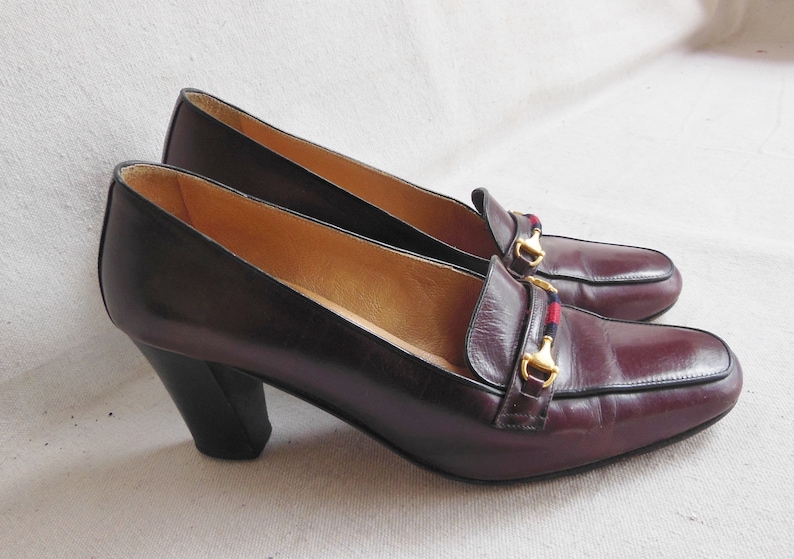 May include: A pair of burgundy leather heeled shoes with a gold buckle and a red and blue stripe detail.