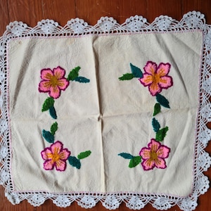 May include: Cream-colored embroidered square textile with four pink and yellow flowers, green leaves, and a white lace border. The flowers have a magenta outline. The textile is likely a decorative item, such as a doily or placemat.
