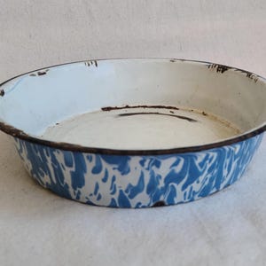 May include: Vintage enamelware bowl with a white interior and a blue and white marbled exterior. The rim is dark brown with signs of wear, adding to its antique charm. A classic piece for kitchen or home decor.