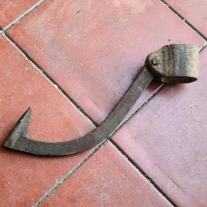 May include: A rusty metal hook with a curved blade and a cylindrical handle. The hook is lying on a red-brown tiled surface. The hook's design suggests it is a tool for butchering or similar tasks.