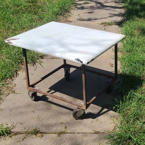 May include: A white metal table with a white top and four wheels. The table is made of metal and has a simple design.
