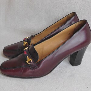 May include: A pair of burgundy leather heeled shoes with gold buckles and a red and black plaid accent. The shoes have a block heel.