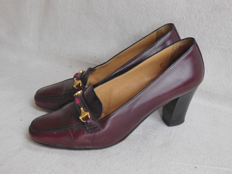 May include: A pair of burgundy leather heeled shoes with gold buckles and a red and black plaid accent. The shoes have a block heel.