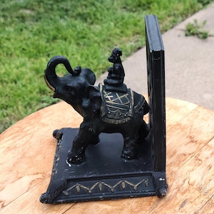 May include: Black elephant bookend with a small monkey figurine riding on its back. The bookend has gold accents and a decorative base.