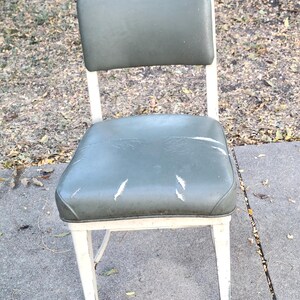 May include: A vintage metal chair featuring a green vinyl seat and backrest. The chair has a white metal frame with signs of wear. The seat shows some scratches. A classic piece of furniture, ideal for a retro aesthetic.