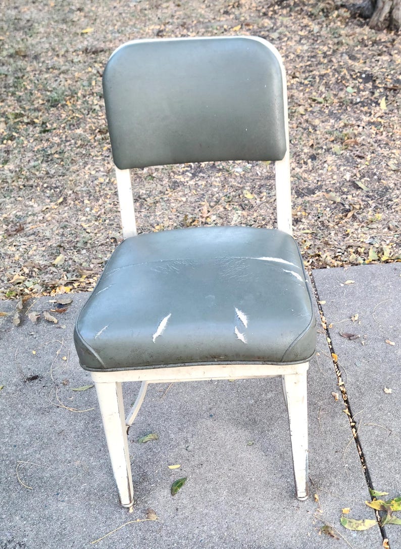 May include: A vintage metal chair featuring a green vinyl seat and backrest. The chair has a white metal frame with signs of wear. The seat shows some scratches. A classic piece of furniture, ideal for a retro aesthetic.