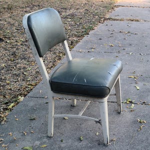 May include: An old metal chair with a dark green padded seat and back. The chair has a white frame showing signs of wear. The chair is positioned on a concrete surface outdoors.