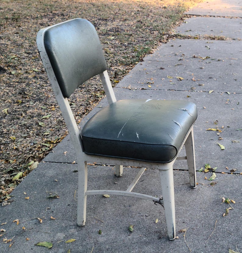 May include: An old metal chair with a dark green padded seat and back. The chair has a white frame showing signs of wear. The chair is positioned on a concrete surface outdoors.