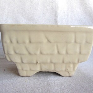 May include: A small, off-white ceramic planter with a brick-like pattern. The planter has four small feet and is designed to hold a small plant.