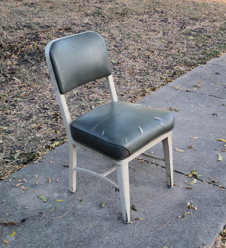 May include: A vintage chair with a dark green seat and backrest. The seat shows signs of wear. The chair has a white metal frame. It is positioned on a concrete surface.