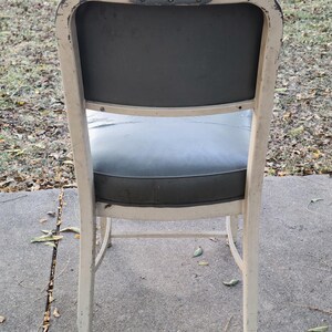 May include: A vintage metal chair with a distressed cream-coloured frame and a dark grey seat and backrest. The chair has a curved back and legs, showing signs of wear and age.