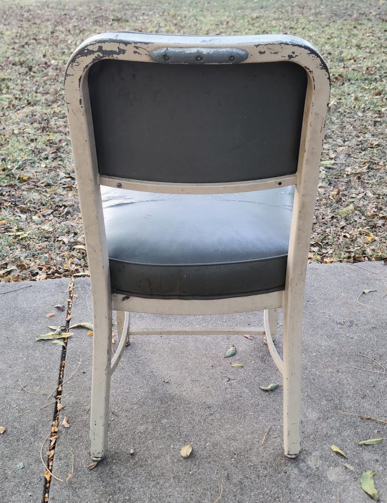 May include: A vintage metal chair with a distressed cream-coloured frame and a dark grey seat and backrest. The chair has a curved back and legs, showing signs of wear and age.