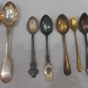 May include: A collection of ten silver, gold, and stainless steel spoons. The spoons have various designs and finishes, including a spoon with a floral design, a spoon with a crown design, and a spoon with a plain finish.