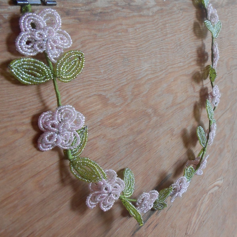 May include: A beaded floral wreath with pink flowers and green leaves. The wreath is made of small beads and is strung together to form a circle.
