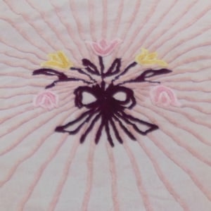 May include: A light pink, textured fabric with a central floral design. The design features a bouquet of embroidered flowers in yellow and pink, tied with a dark purple bow. The background has radiating lines.