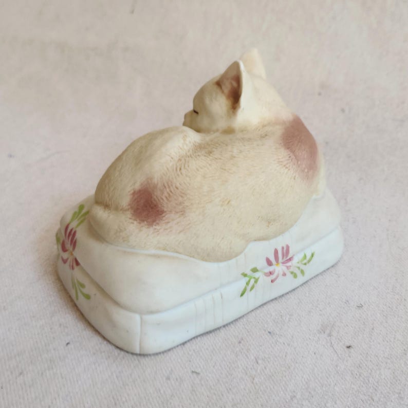 May include: A ceramic figurine of a sleeping cat, cream-colored with pink spots, resting on a white pillow. The pillow is decorated with pink and green floral designs. The cat is curled up, facing away from the viewer.