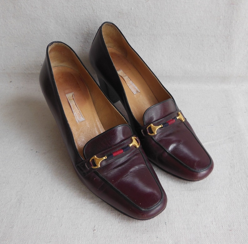 May include: A pair of burgundy leather heeled loafers with gold horse bit accents and a red and blue stripe detail.