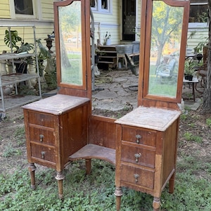 May include: A wooden vanity table featuring a large central mirror flanked by two adjustable side mirrors. The piece includes two sets of three stacked drawers with small metal pulls, supported by four legs, with a central open space for seating.