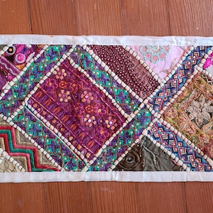 May include: A colorful, patchwork table runner with a white border. The runner features a variety of patterned fabrics in shades of purple, pink, teal, and gold, arranged in diamond shapes. The design includes embroidered details and decorative stitching.