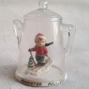 May include: A clear plastic snow globe shaped like a coffee pot with a small figurine of a child skiing inside. The base of the snow globe has the words "Manistee Michigan" printed on it.